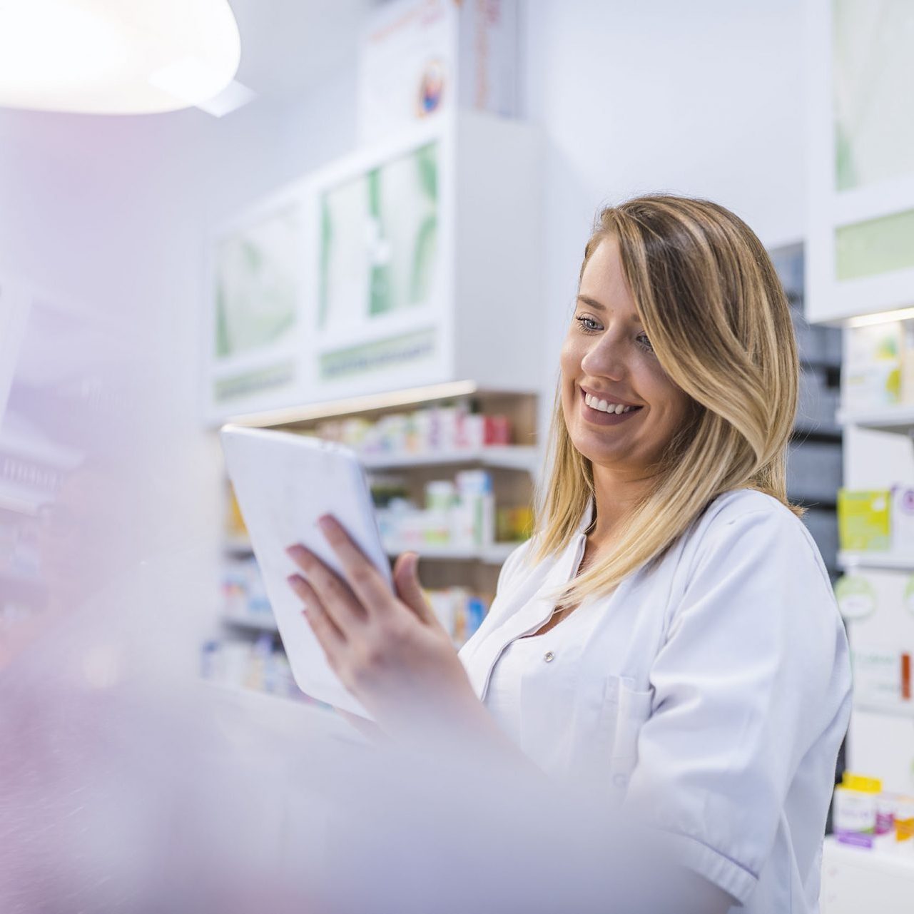 Pharmacist working with a tablet computer in the pharmacy holding it in her hand while reading information. Cheerful happy pharmacist chemist woman working in pharmacy drugstore with tablet computer