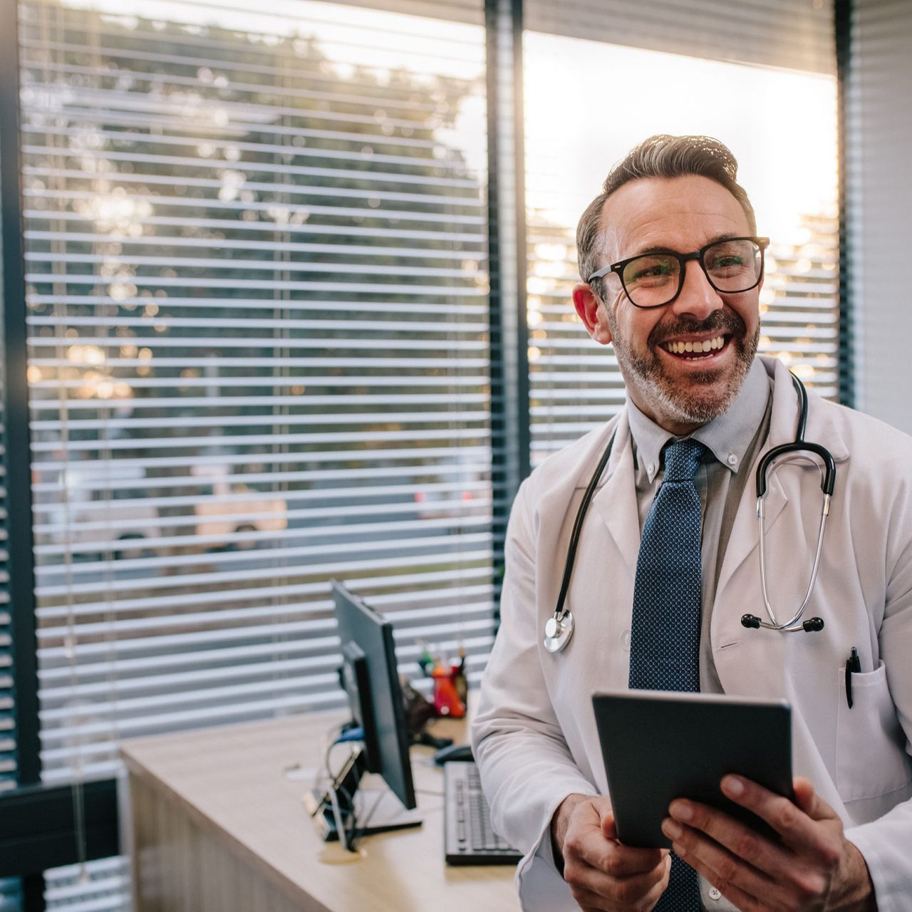 Smiling mature male doctor with digital tablet in his office. Friendly medical professional with tablet computer in clinic.